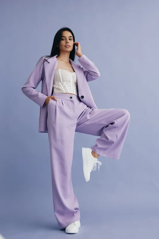 young woman in lilac suit posing confidently against plain purple background wearing white sneakers and lace top, stylish modern outfit, casual fashion look