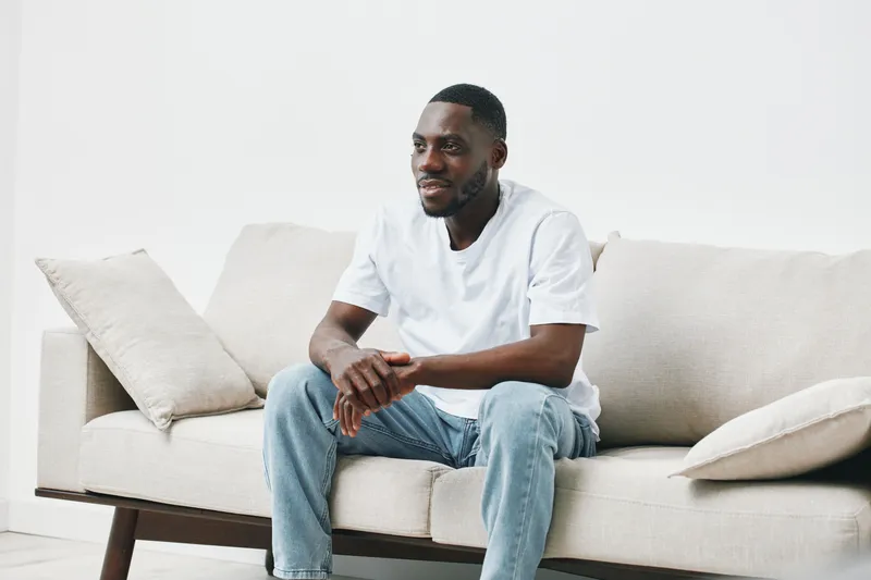 Relaxed young African American man in casual white t shirt sitting on a light sofa, expressing happiness in a minimalist indoor setting