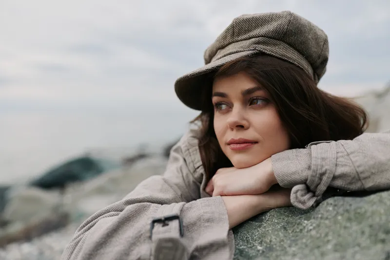 Young woman in casual beige clothing and hat leans on a rock outdoors, looking away thoughtfully. Soft natural light highlights her relaxed, contemplative mood in a serene environment.