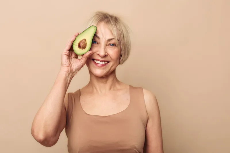 Happy senior woman with short hair holds fresh avocado half near eye, smiling in beige tank top. Concept of healthy lifestyle, nutrition, natural food, and positive aging.
