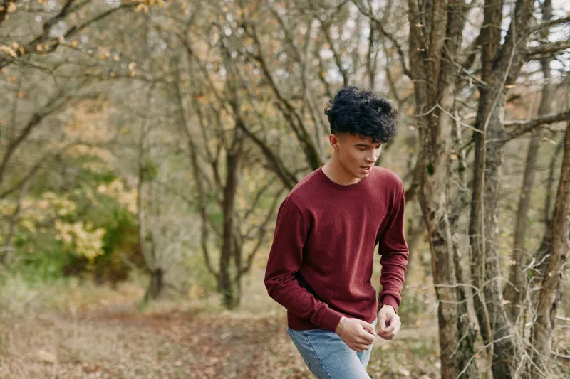 An authentic, candid portrait of a young man in a burgundy sweater amid an autumn forest, conveying genuine mood, natural posture, and everyday style.