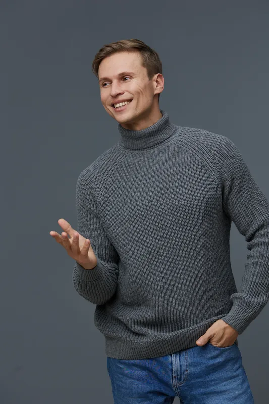 Smiling young man wearing a gray turtleneck sweater and jeans, standing against a plain gray background in a studio setting, casually gesturing with one hand, cheerful and approachable personality. Pe