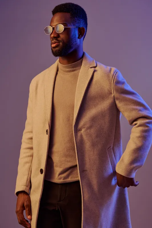 Stylish African American man in a light coat and sunglasses, showcasing confidence against a vibrant purple background Stylish fashion concept for modern elegance