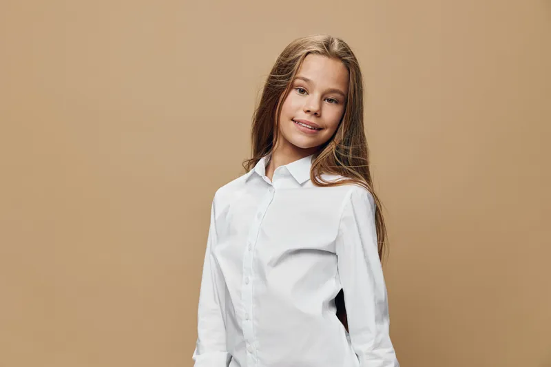 Girl smiling and posing with long hair wearing white shirt against beige background. Child with natural expression and casual style in studio portrait with soft lighting and simplicity.