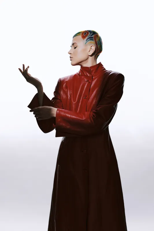 Woman with colorful short hair wearing a shiny red leather coat posing gracefully against a light background, showcasing contemporary fashion and artistic style.