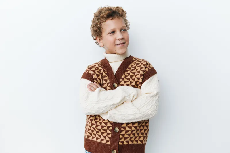 A young boy stands with arms crossed, wearing a brown and cream geometric knit vest over a white turtleneck sweater, exuding confident, playful charm against a clean neutral backdrop.