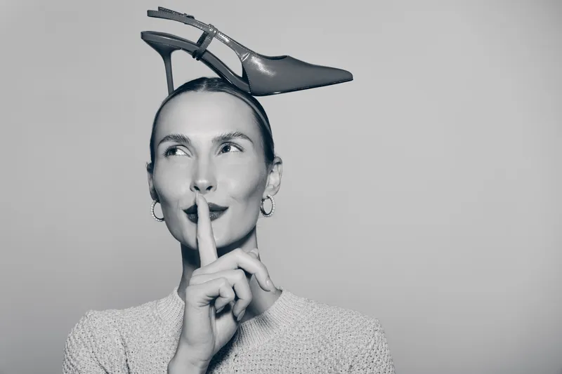 White woman with fair skin holds finger to lips in shushing gesture with high heel shoe on head in studio.