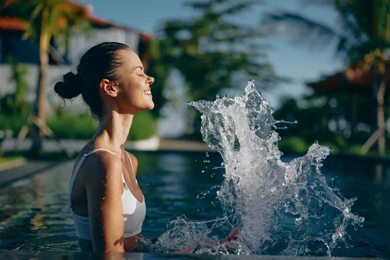 Smiling woman in a white swimsuit enjoys a splash by the pool on a sunny day, lifestyle moment, wellness vibe, natural light emphasizes a cheerful refreshing mood.