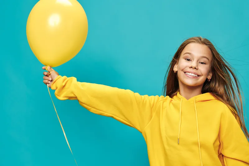 Smiling girl in yellow hoodie holding yellow balloon with outstretched arm on blue background, happy child portrait with long hair and casual clothing for celebration or fun.
