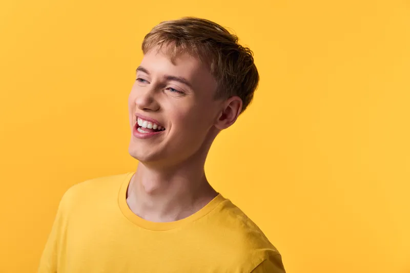 Smiling man, yellow shirt, bright background, happy mood, joyful expression, cheerful demeanor in a studio setting with daylight lighting and warm tones, conveying upbeat energy and friendliness