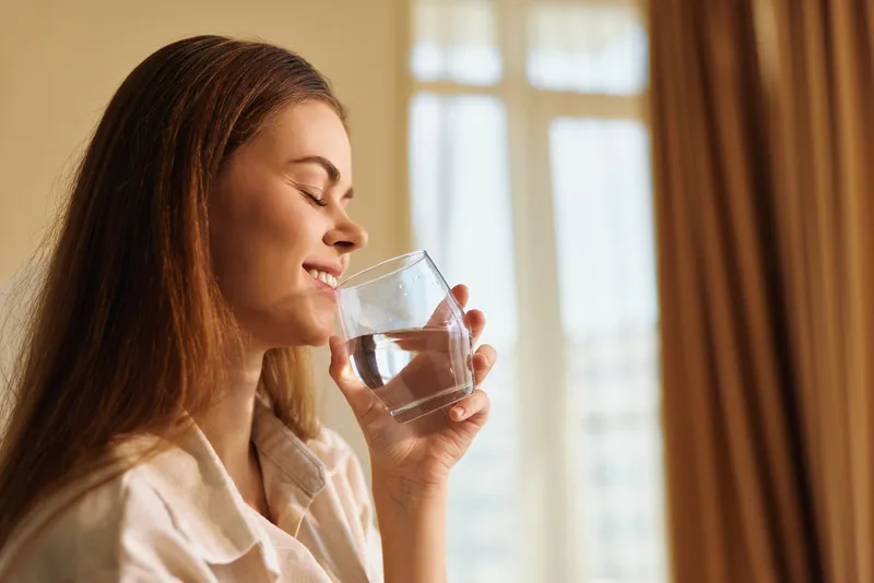 Woman drinking water from glass at home in morning, relaxed and healthy routine, closeup profile with soft natural light, hydration and wellness for daily self care.
