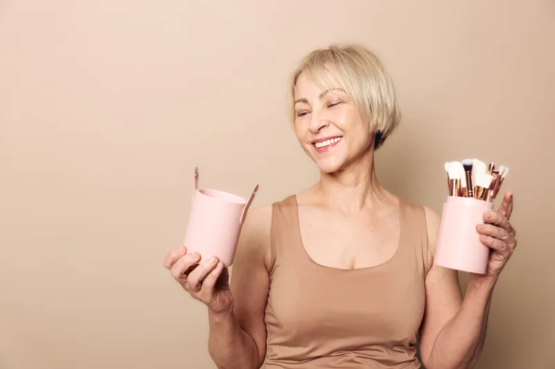 Smiling mature woman with short blonde hair holds pink containers with makeup brushes on beige background. Concept of beauty routine, cosmetics tools, self-care, and joyful lifestyle.