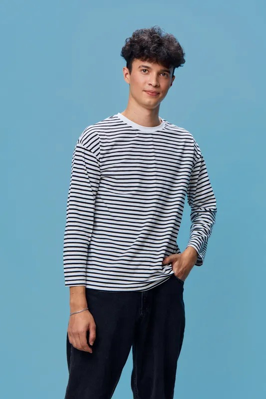 young man in striped shirt standing with casual pose against a solid blue background, dressed in dark pants with a relaxed expression and curly hair