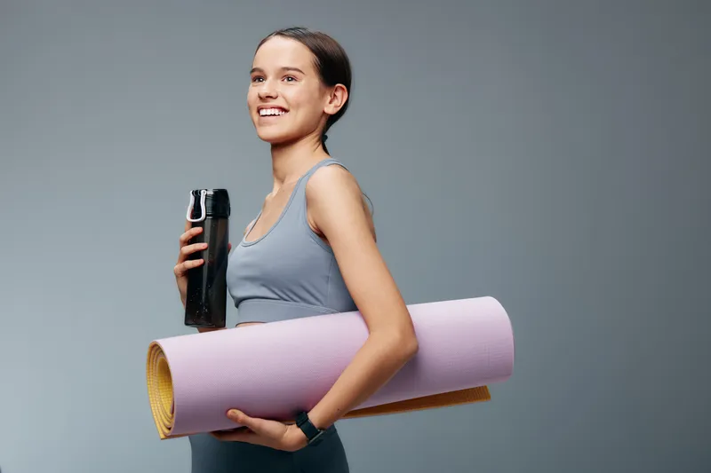 Female fitness enthusiast holding a yoga mat and a water bottle, ready for training. Casual athletic wear, neutral background, focused and confident.