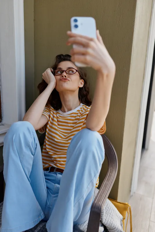 Selfie of a woman wearing a striped shirt and light jeans, seated on a balcony, holding a smartphone. Casual home life, relaxed mood, natural light, candid moment.