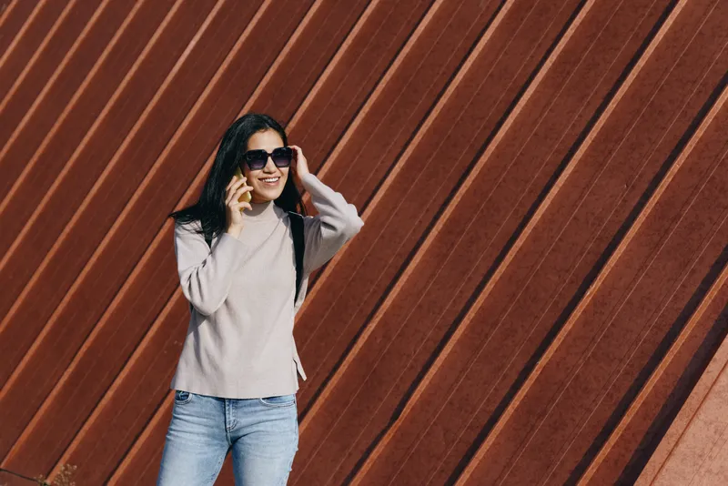 Fashionable woman wearing sunglasses and a light sweater stands against a diagonal rusted wall, showcasing outdoor casual chic and urban street style for contemporary fashion photography, lifestyle
