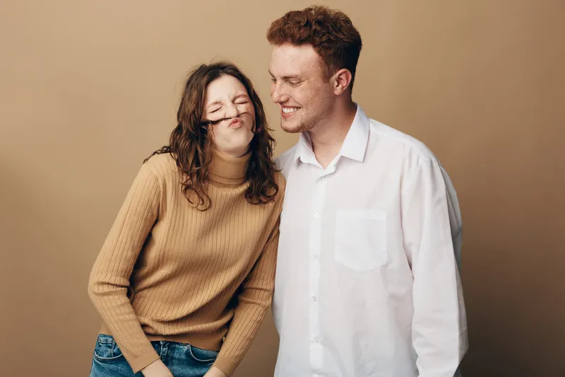 Young couple having fun together, woman making a funny face with hair mustache, man smiling in casual clothes on beige background. Concept of friendship, joy, and playful moments.