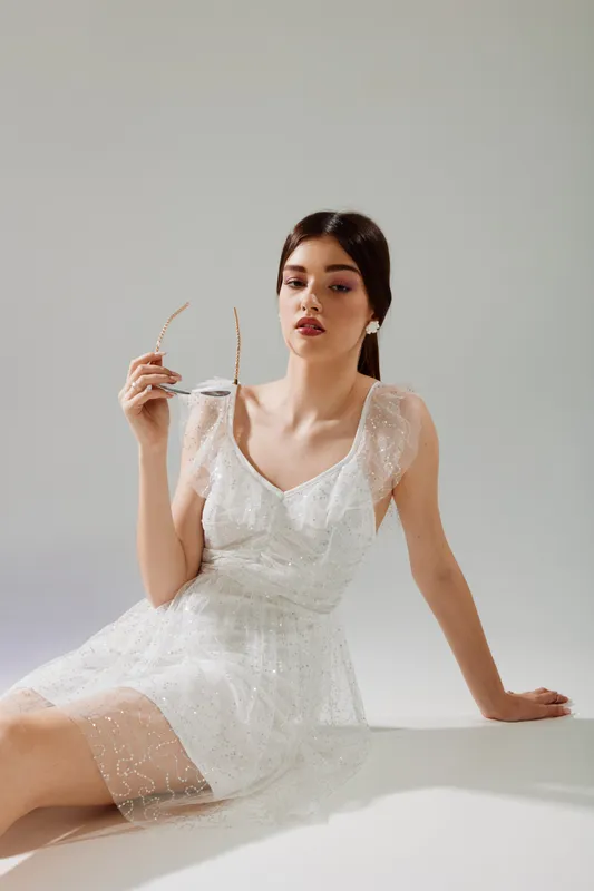 Elegant woman beauty fashion model in a delicate white dress holding sunglasses sitting on a white surface. Studio shot with soft lighting and minimalistic background.
