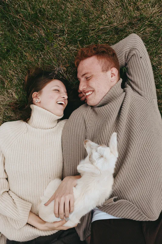 Young couple lying on grass wearing knitted sweaters enjoy a playful moment together while holding a small white baby goat in a natural outdoor setting.