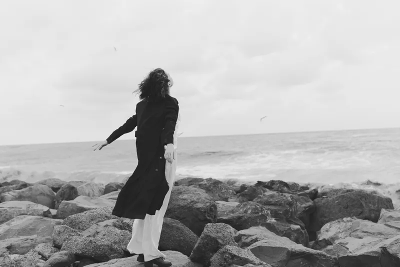 Woman in long coat stands on rocky shore facing the sea, wind in her hair, monochrome scene capturing solitude, coast, horizon, and movement along the water