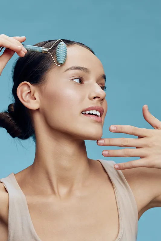 Young woman using a facial roller for skincare, showcasing glowing skin and a relaxed expression, against a soft blue backdrop for beauty and wellness purposes