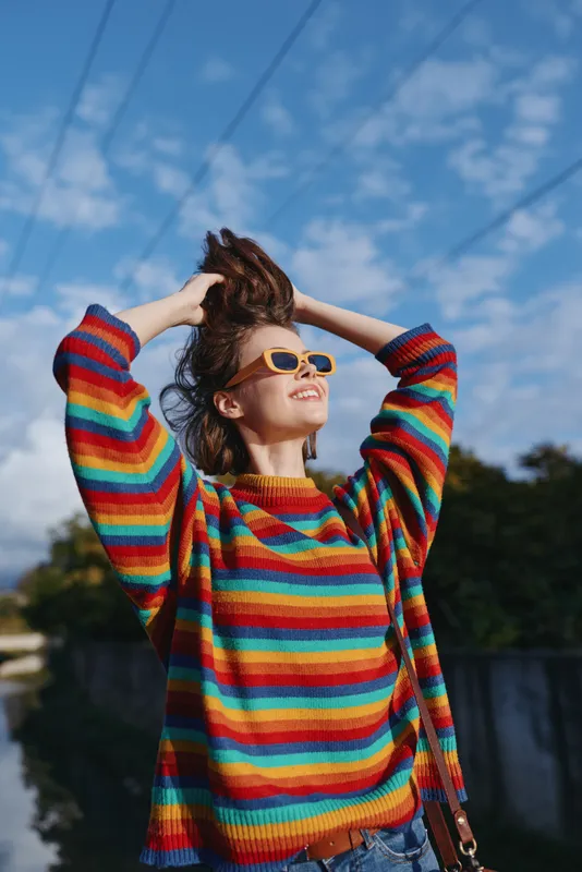 Young woman in her 20s wearing a colorful striped sweater and yellow sunglasses, smiling outdoors under blue sky with hands in hair, casual outfit and denim jeans on a sunny riverside travel