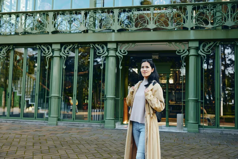 Woman in a long beige coat stands near an ornate storefront with green ironwork, urban street backdrop, casual chic style, daylight photography mood, fashion moment, street photography vibe