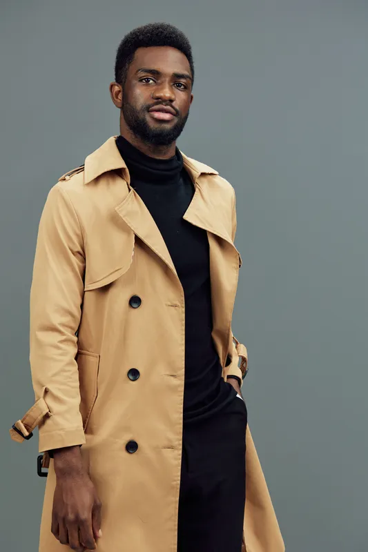 Stylish African man in beige trench coat exuding confidence against a gray background, projecting a modern fashion vibe perfect for seasonal trends