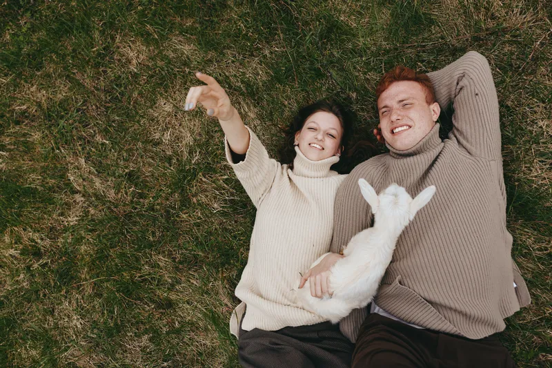 Couple lifestyle scene shows a smiling pair lying on grass, taking a selfie with a friendly goat kid, enjoying relaxed affection and countryside charm in a sunny outdoor moment.