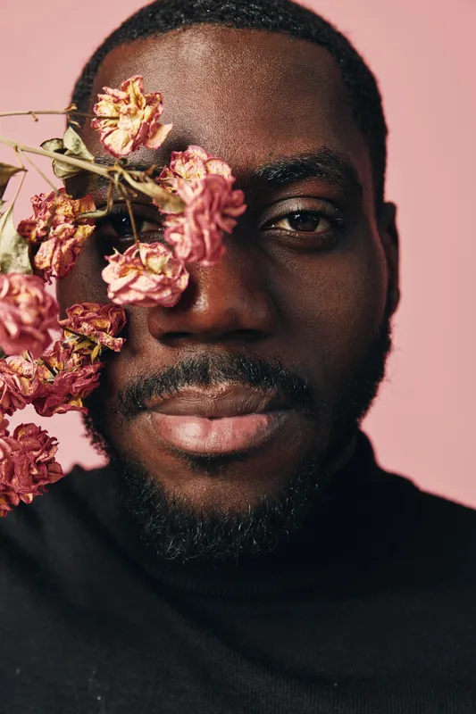 Creative portrait of African man with dried pink flowers on face in conceptual visual style. Closeup, black turtleneck, natural skin texture, artistic expression and soft pink background.