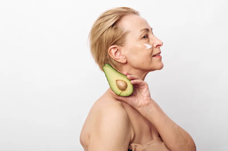Mature woman holds avocado slice near neck, applying natural face cream on cheek. Concept of healthy skincare, organic beauty, anti aging routine, and wellness in minimalist studio setting.