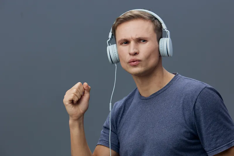 Young man wearing blue t-shirt and large headphones, expressing excitement or enthusiasm, clenched fist gesture, studio shot with plain gray background, lifestyle concept, music enjoyment.