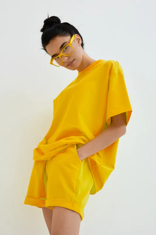 young woman wearing bright yellow casual outfit and matching glasses, standing against white background with relaxed and stylish pose, modern fashion look