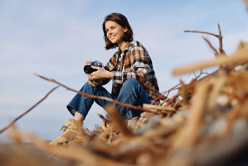 Woman outdoors on a sandy dune, holding a camera, wearing a plaid shirt and jeans, focused on nature photography under a clear sky with driftwood nearby.