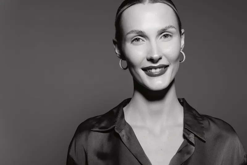 Portrait of a smiling Caucasian woman with light skin and slicked-back hair wearing hoop earrings and a shiny shirt. Studio lighting highlights her natural beauty and confident expression.