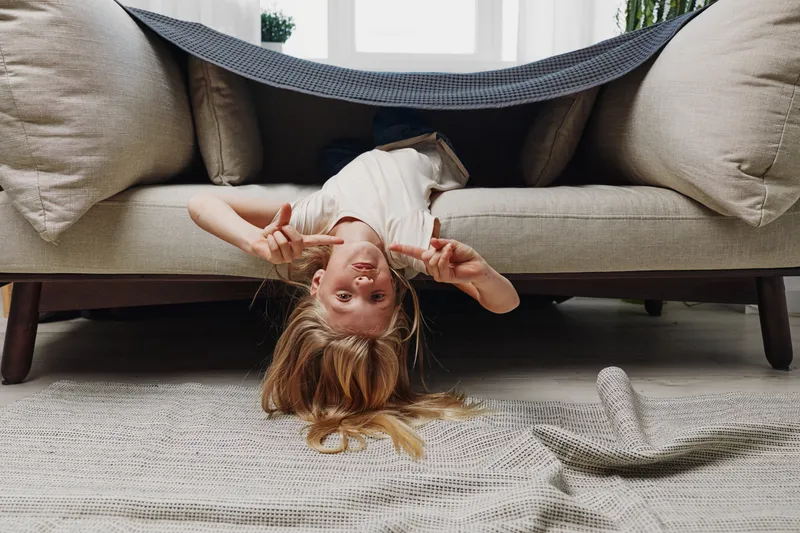 Playful child with blonde hair upside down under couch blanket fort shows peace signs and smiles in cozy living room. Concept of childhood fun and imagination.