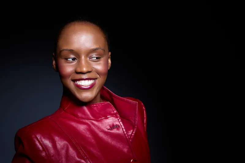 Smiling African woman with dark skin wearing a red leather jacket looks to the side. Studio portrait with black background. Concept for fashion, confidence, and beauty.