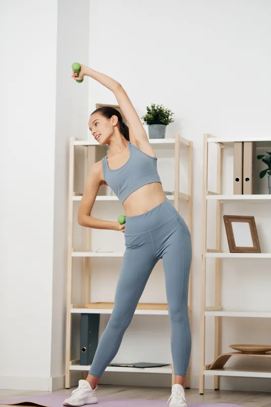 Woman fitness in a bright living room performing a side stretch with dumbbells for balance and tone, showcasing a focused workout routine in a modern home gym setting.