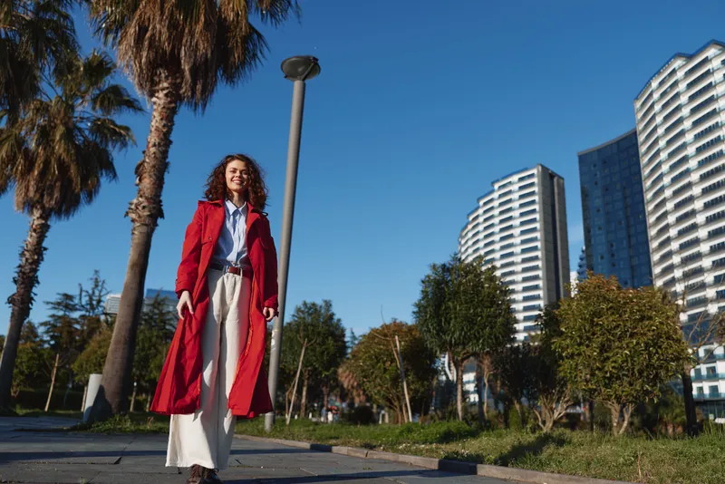 A stylish woman walks outdoors in a bright red coat along a city park, with palm trees and modern buildings in the background. A bold street style concept for contemporary fashion.