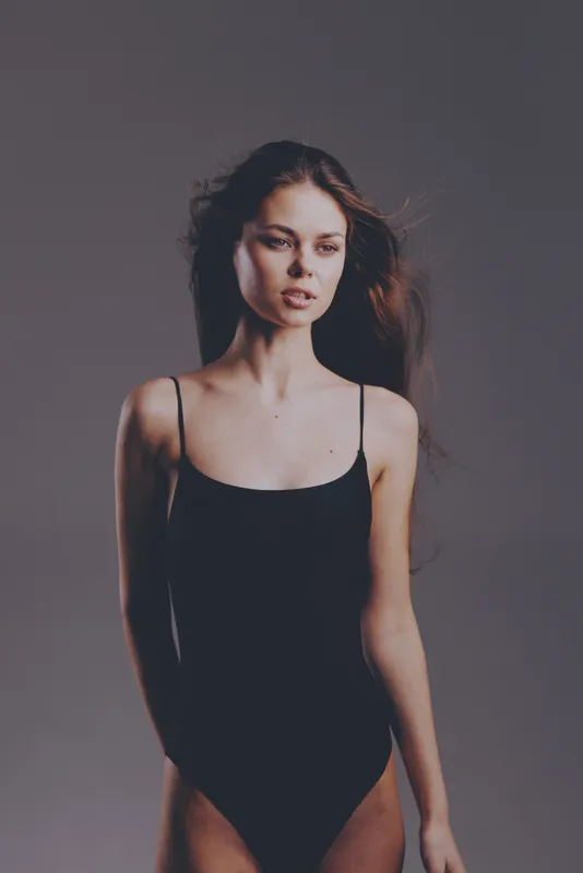 Young woman in black bodysuit posing with serious expression against neutral background