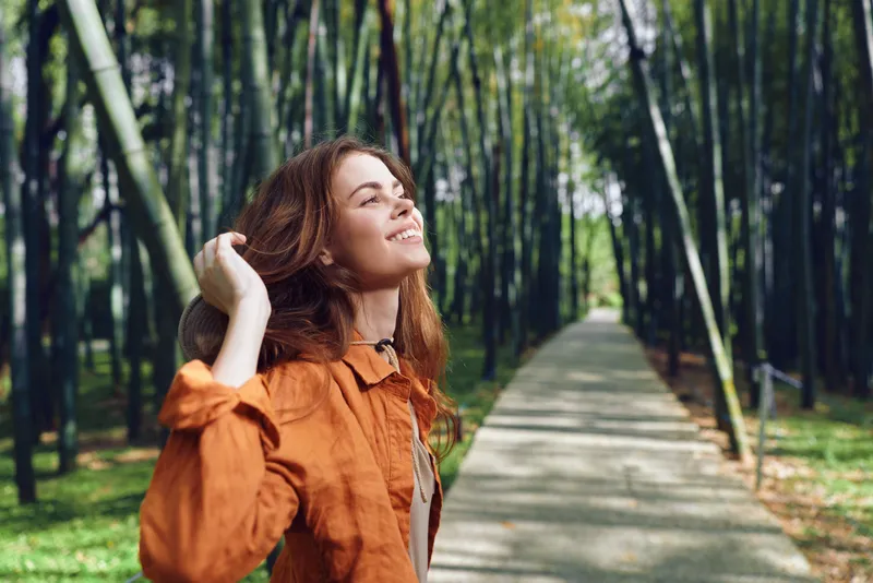 Woman smiling on a sunlit bamboo forest path wearing a hat and orange jacket, eyes closed and head tilted back, enjoying nature, fresh air and a serene walking portrait for relaxation.