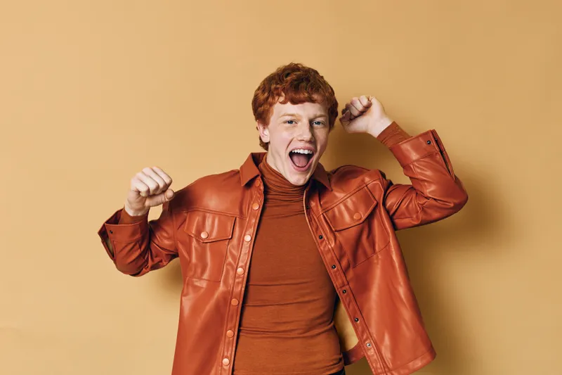 Young man with red hair wearing brown leather jacket and sweater, smiling and raising fists in excitement against beige background. Casual style and energetic mood.