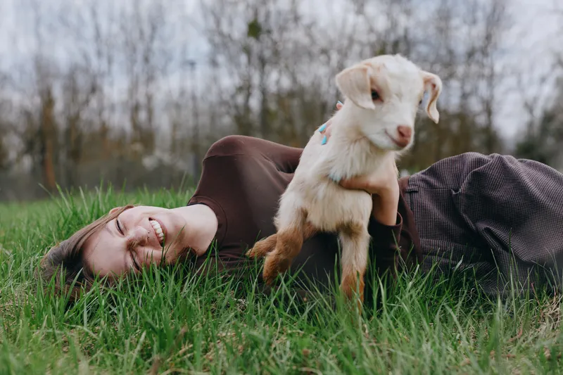 A relaxed woman lies on lush grass outdoors, cradling a smiling goat kid in her arms. A candid lifestyle moment highlighting bonding, care, and gentle play.
