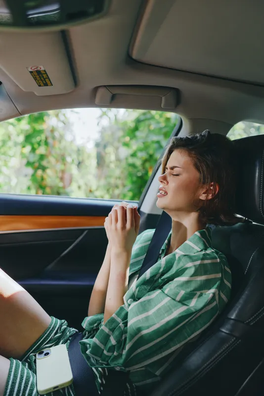 Young woman sits in a car seat wearing a striped green shirt and a seatbelt, resting with eyes closed as light fills the cabin.