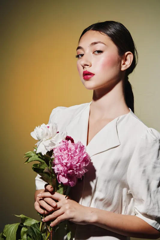Young woman with a sleek hairstyle and bold makeup holds pink and white flowers. She wears a white outfit in soft warm studio lighting. Concept for beauty creative fashion photography.