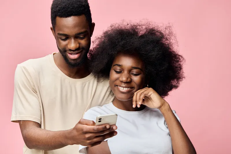 Happy young African couple smiling together while looking at a smartphone against a soft pink background, showcasing joy and togetherness