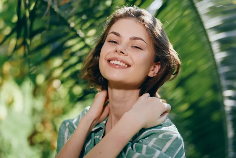 Bright portrait of a smiling woman outdoors, bathed in sunlight with lush green foliage in the background.