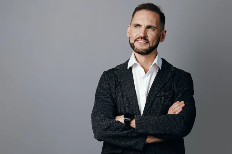 A poised male business professional stands with crossed arms against an isolated colored background, conveying confidence, focus, and calm determination in a studio portrait.