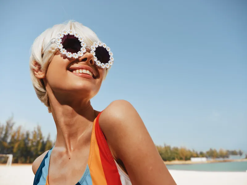 A stylish woman on a sunny beach wears sparkling sunglasses and a vibrant swimsuit, smiling toward the sky as sea and shoreline blur softly in the background.