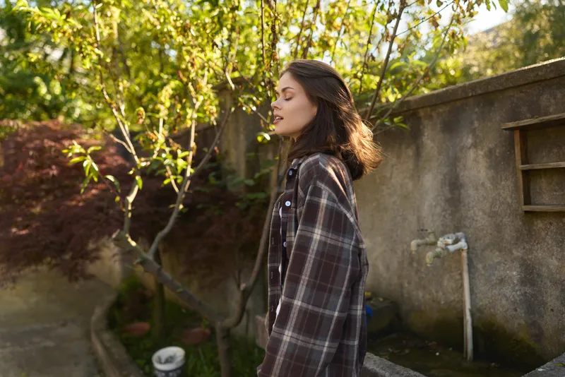 A woman in a plaid jacket enjoys a calm moment outdoors, her profile framed by sunlit greenery. A lifestyle concept centered on everyday simplicity, personal style, and outdoor exploration